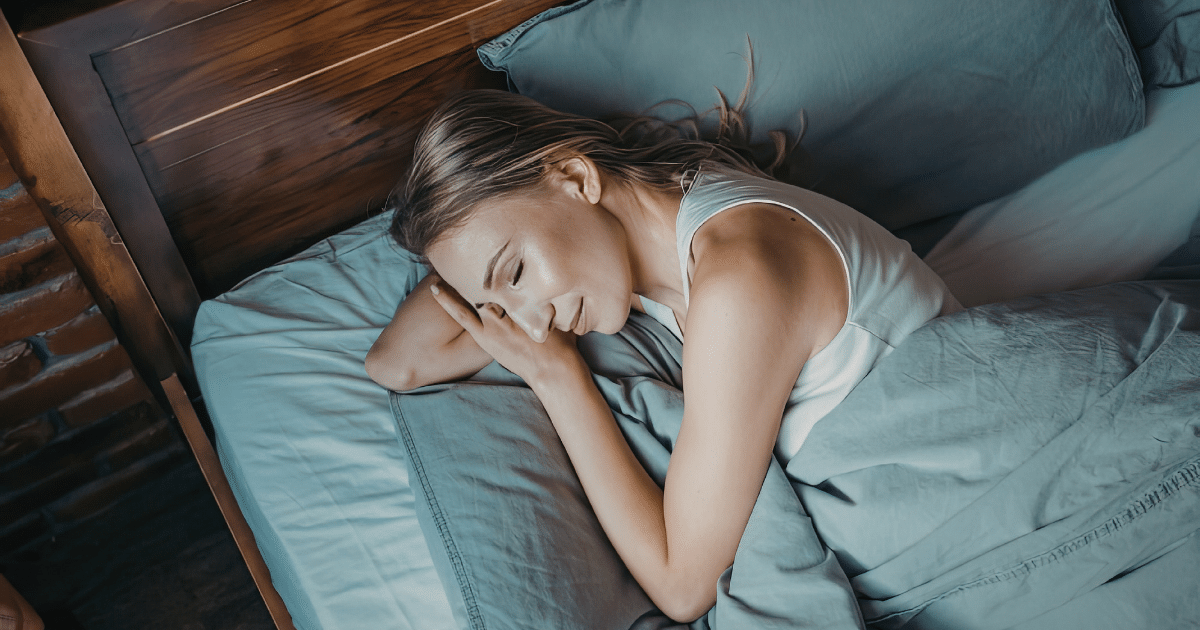 A high-angle shot of a blonde woman sleeping peacefully in a bed. She is wearing a white tank top, lying on her side with a gentle smile on her face, and resting her hands near her pillow.