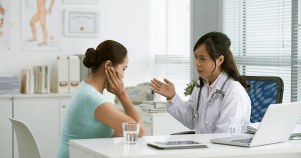 A medical consultation scene where a female doctor in a white lab coat with a stethoscope is talking and gesturing with her hand toward a female patient who sits across from her, holding her hand up to her neck or cheek area.