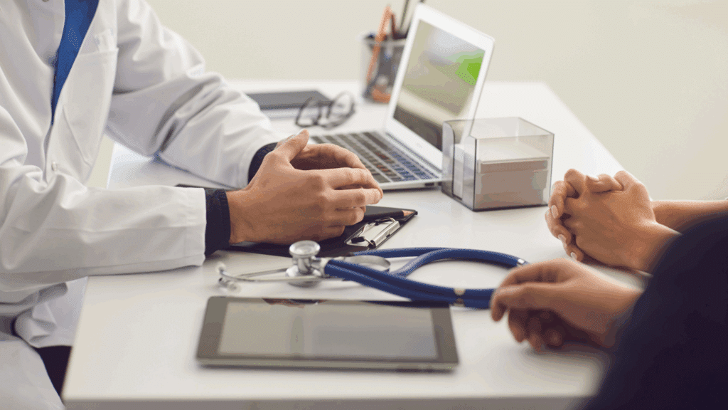 A doctor in a white lab coat talking to a seated patient; only the doctor’s arms and coat are visible, with their face out of frame.
