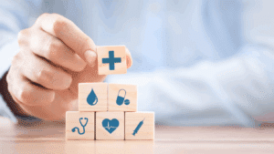 A hand is placing a white cube with a medical cross icon on top of a pyramid of similar cubes. Each cube features different healthcare-related icons, including a stethoscope, pills, ambulance, heart rate, and a phone.