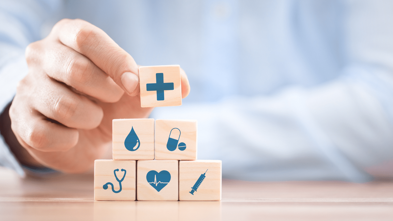 A hand is placing a white cube with a medical cross icon on top of a pyramid of similar cubes. Each cube features different healthcare-related icons, including a stethoscope, pills, ambulance, heart rate, and a phone.