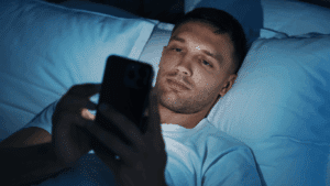 A man lying in bed at night, using electronic gadgets.