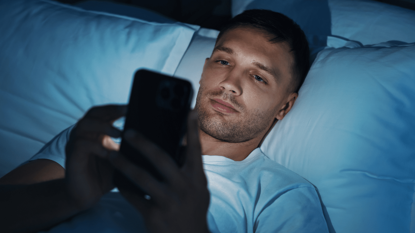 A man lying in bed at night, using electronic gadgets.