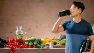 Man in a blue shirt drinking from a shaker bottle while holding a supplement container, standing beside a table with fresh fruits, vegetables and supplements.