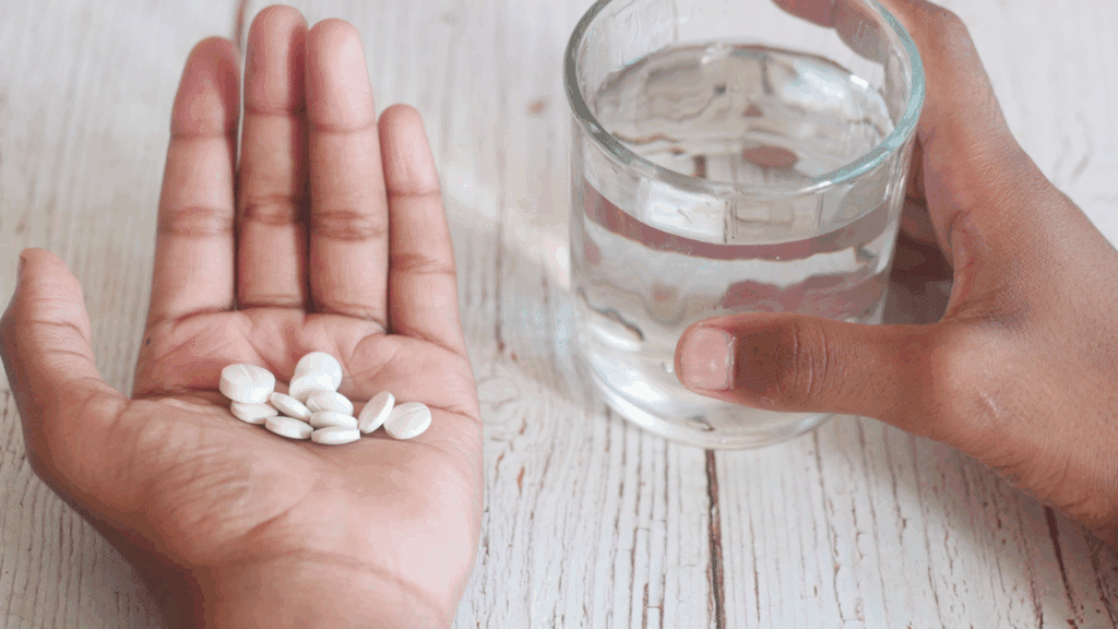 A hand holding several supplement pills beside a glass of water.