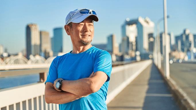 Middle-aged Asian man in a blue athletic shirt and a baseball cap standing on an elevated walkway with a city skyline in the background, arms crossed.