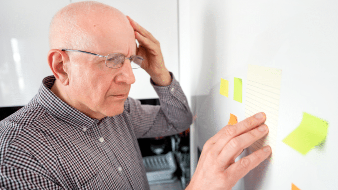 Bald, older man wearing glasses looking closely at colorful sticky notes on a white board, with his hand on his head.