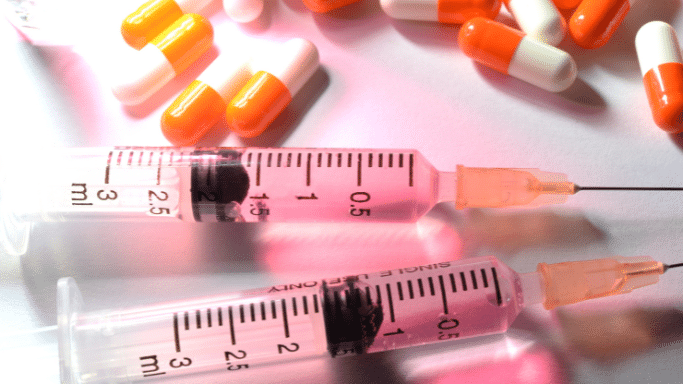 Close-up of two syringes containing pink liquid, with white and orange pill capsules scattered in the background.