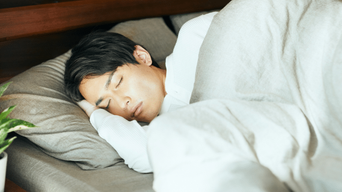 A young man with dark hair is peacefully sleeping on his side under a light-colored duvet in a cozy bedroom.