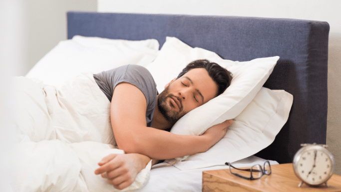 A man with a beard is sound asleep on his side, under white bedding, with an alarm clock and glasses on a nightstand nearby.