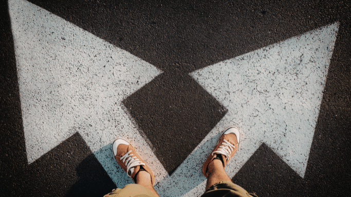 A top-down perspective of a personโs feet standing on dark asphalt. Painted on the ground are two large, white arrows pointing in opposite directions (left and right).