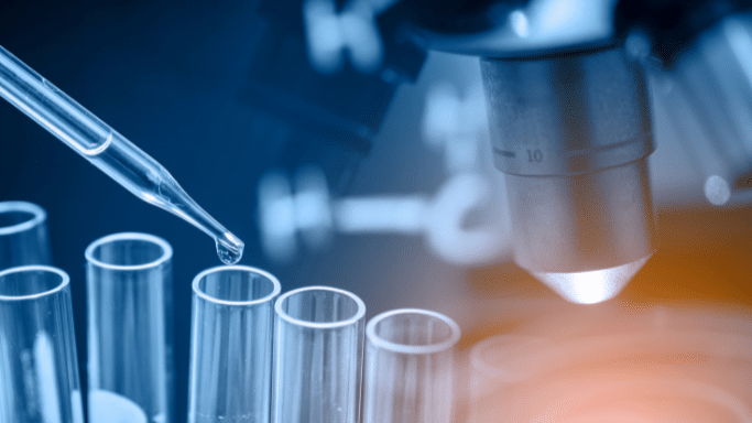 A stylized, blue-toned close-up of laboratory equipment. A glass pipette is shown suspended over a row of test tubes, with a single drop of liquid about to fall. A microscope lens is positioned in the background on the right, illuminated by a warm orange glow.