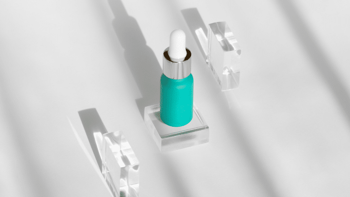 A top-down, minimalist shot of a small, teal-colored nasal bottle sitting on a clear glass block. Two additional clear glass blocks are placed nearby on a white surface with sharp diagonal shadows.
