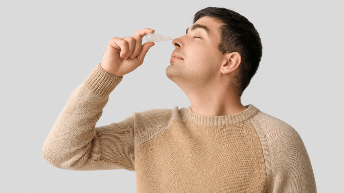 A side-profile view of a man in a tan sweater with his eyes closed, holding a small white dropper bottle up to his nose to administer nasal drops.