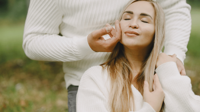 Close-up of a blonde woman with her eyes closed, smiling slightly, as a man's hand gently touches her cheek. They are both wearing white ribbed sweaters and are outdoors.
