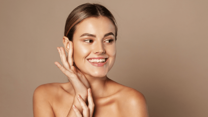 A young woman with her hair pulled back, smiling brightly and looking off-camera, gently touching her cheek and neck with both hands.
