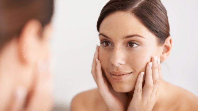 A young woman with dark hair looks intently at her reflection in a mirror, holding her hands to her cheeks.