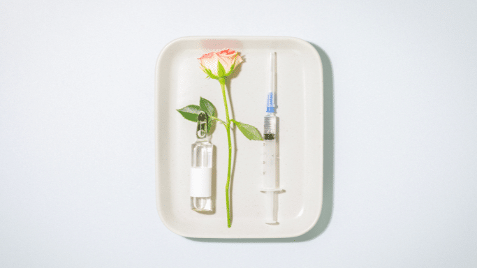 A still life image on a white tray featuring a single pink and white rose, a glass ampoule with a white label, and a syringe with a clear liquid.