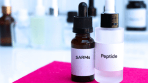 Two small dropper bottles, one amber-colored labeled "SARMs" and one clear/frosted labeled "Peptide," sitting on a bright pink surface in a laboratory or scientific setting with other blurred bottles in the background.