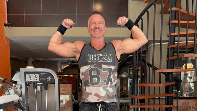 The man with the mohawk poses in a gym, wearing a camouflage "IBIZA 87" tank top and wrist wraps. He is performing a double bicep flex and smiling, with a spiral staircase visible in the background.