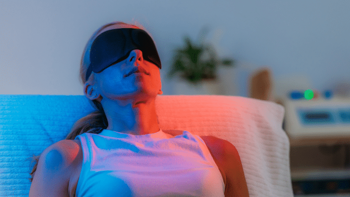 A person wearing a black sleep mask reclines on a white cushioned chair. The scene is lit with a dual-tone lighting effect, casting cool blue light on one side of their body and warm red light on the other.