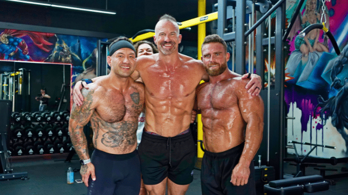 Three muscular, shirtless men pose together in a vibrant gym. The man in the center has his arms around the other two; all are smiling and appear sweaty from a workout. Colorful superhero-themed murals decorate the background walls.