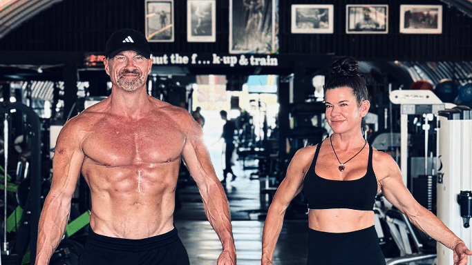 A shirtless, muscular man with a black baseball cap and a muscular woman in a black sports bra stand side-by-side in a well-equipped gym. A