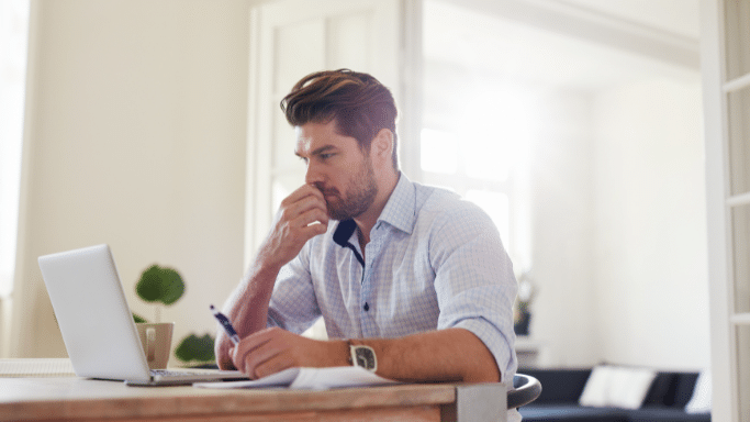 A man in a light blue checkered shirt sits at a wooden table, looking intently at his laptop screen. He has a pen in one hand and is resting his chin on his other hand, looking deeply concentrated on his work.