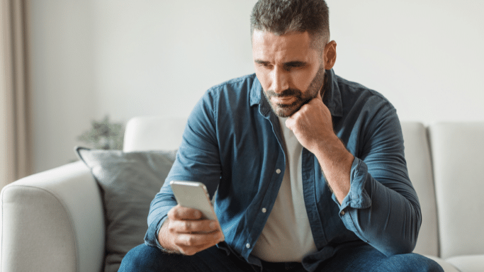 A man sitting on a light-colored sofa, leaning his chin on his hand while looking down intently at his smartphone. He is wearing a blue button-up shirt over a white t-shirt.