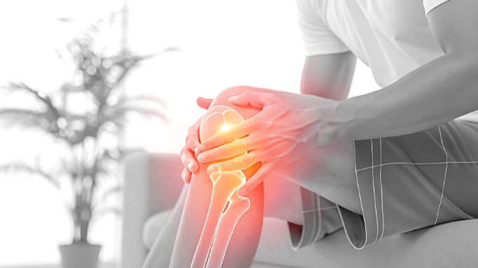 A medical illustration of a person sitting and clutching their knee. A glowing red and orange heat map is overlaid on the knee joint and surrounding leg bones to indicate intense pain or inflammation.