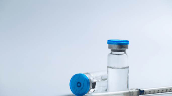Two medical glass vials with bright blue caps containing clear liquid sit on a light blue surface next to a plastic syringe. One vial stands upright while the other lies on its side.