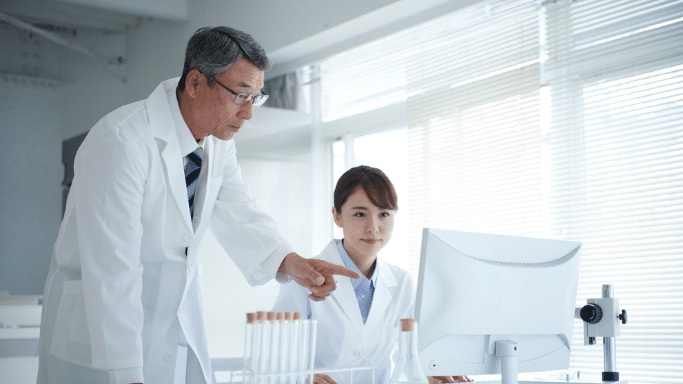 Two scientists in white lab coats, an older man and a younger woman, working in a bright, modern laboratory. The man is leaning over and pointing at a computer monitor while the woman looks on, with lab equipment like test tubes and a microscope visible in the foreground.