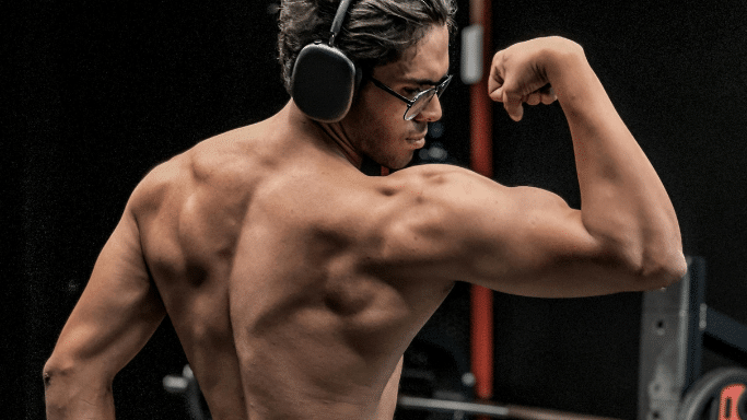 A shirtless man wearing glasses and large over-ear headphones seen from behind. He is flexing his arm, highlighting the well-defined muscles of his back, shoulders, and bicep.