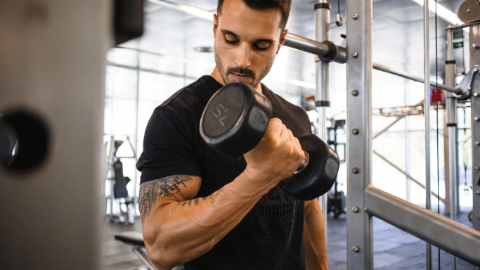 A man with arm tattoos performing a bicep curl with a large dumbbell in a gym. He is focused on his form, with exercise machines and gym equipment visible in the blurred background.