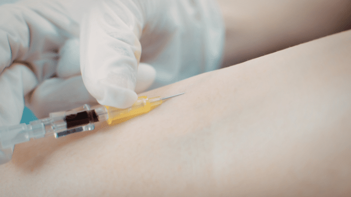 A close-up shot of a gloved hand holding a needle as it is being inserted into a person's skin for a blood draw or injection.