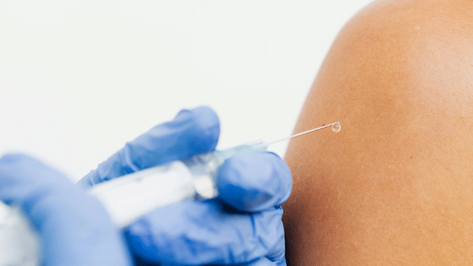 An extreme close-up of a syringe with a drop of clear liquid on the needle tip, positioned near a person's shoulder.