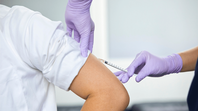 A healthcare professional wearing purple gloves administers an injection into the upper arm of a patient wearing a white shirt.