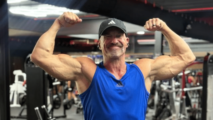 A close-up of a muscular man in a gym, wearing a blue Adidas tank top and a black baseball cap. He is smiling while performing a front double-biceps pose, showcasing his arm and shoulder definition.