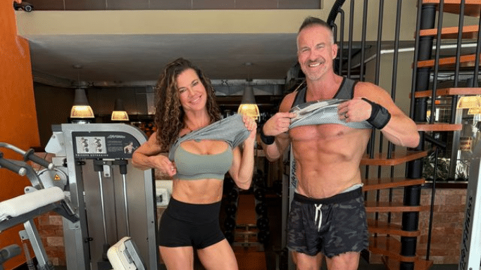 A man and woman posing together in a gym setting, hey are both lifting the bottom of their shirts to reveal their toned abdominal muscles.