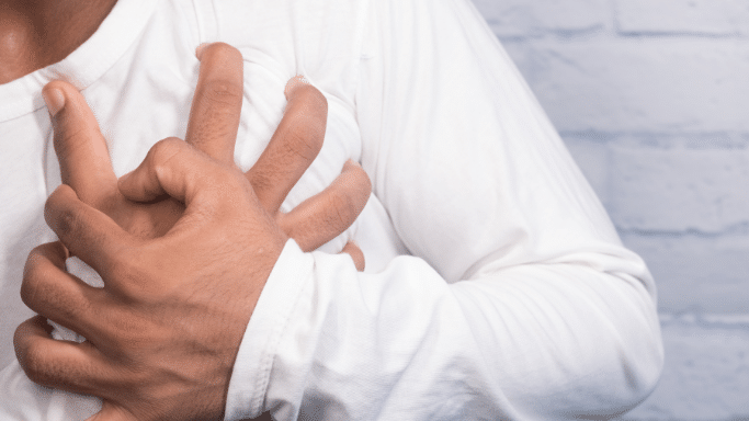 A close-up shot of a person wearing a white long-sleeved shirt, clutching their chest with both hands in a gesture indicating pain or a medical emergency, such as a heart attack. The background is a light-colored brick wall.