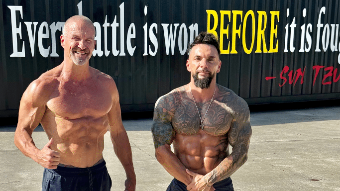Two muscular, shirtless men standing side-by-side outdoors. The man on the left is smiling and giving a thumbs up; the man on the right has extensive tattoos on his chest and arms.