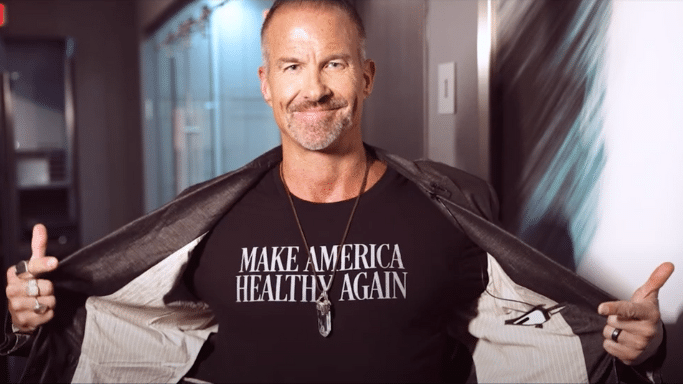 A smiling middle-aged man with short hair and a goatee, wearing multiple rings and a crystal pendant necklace. He is holding open a dark blazer to reveal a black t-shirt that reads "MAKE AMERICA HEALTHY AGAIN" in white, capitalized serif font.