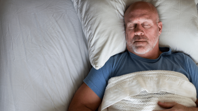 An overhead shot of a middle-aged, bald man with a grey beard sleeping peacefully in a bed. He is tucked under a white patterned blanket and resting his head on a white pillow.