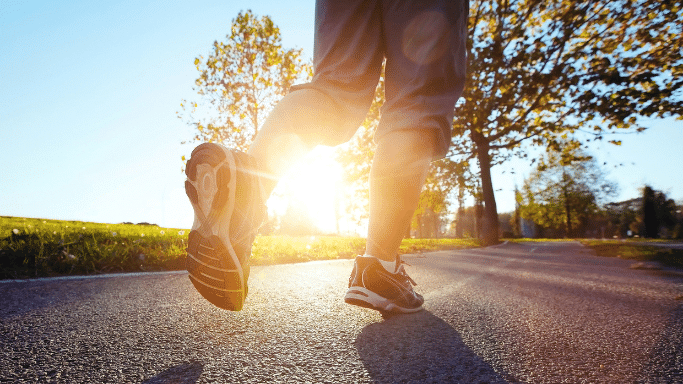 A low-angle, close-up shot of a runner's feet on an asphalt path during a bright sunset or sunrise. The sun shines low between the runner's legs, creating a strong glow and long shadows on the ground.