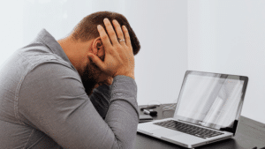 A man in a grey button-down shirt sits at a desk, leaning forward with his head buried in his hands in a gesture of stress or exhaustion. An open laptop is on the desk in front of him.