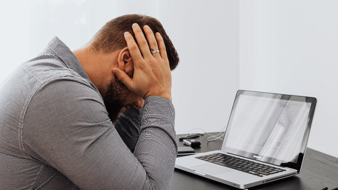 A man in a grey button-down shirt sits at a desk, leaning forward with his head buried in his hands in a gesture of stress or exhaustion. An open laptop is on the desk in front of him.