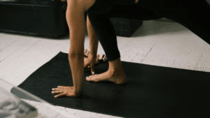A close-up, low-angle shot of a person practicing yoga on a black mat. The focus is on their hands and one foot firmly planted on the mat in a lunge-like position.