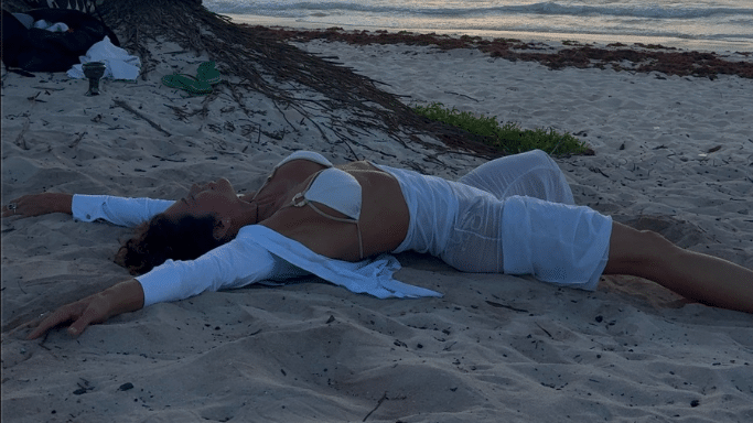 A woman lies on her back on a sandy beach at dusk, her arms spread wide. She is wearing a white bikini and a sheer white cover-up.