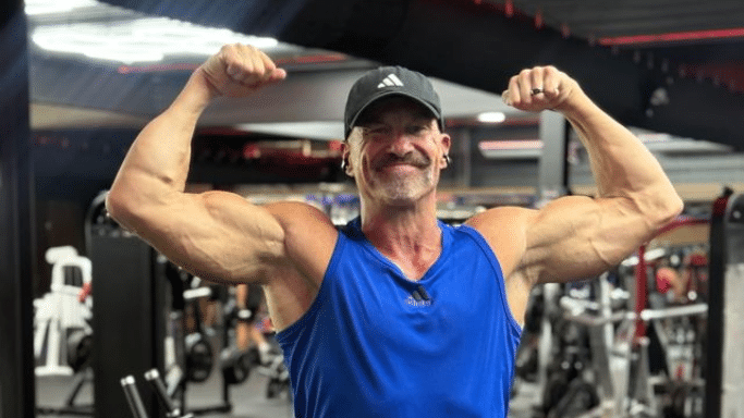 A fit, older man with a mustache and a black baseball cap smiling and flexing both of his muscular biceps in a gym setting. He is wearing a blue Adidas tank top.