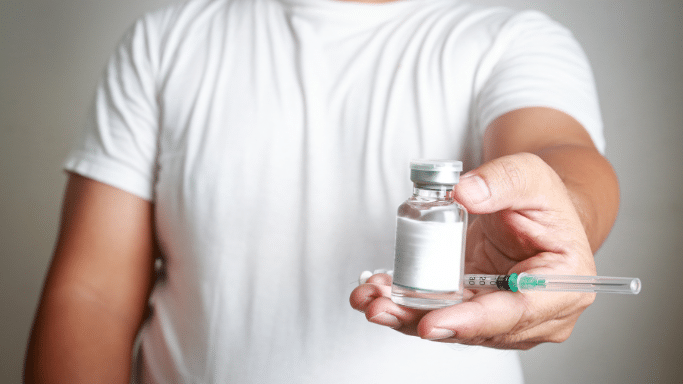 A person in a white t-shirt holding a small glass medication vial in the foreground, with a medical syringe resting across their palm. The background is a neutral, light gray.